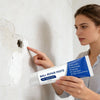 A woman points to a small hole in a wall while holding a tube of Wall Repair Paste for drywall repair.