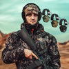 A man wearing professional noise reduction ear muffs and a tactical jacket holds a rifle in an outdoor, arid landscape.