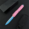 A pink and blue tactical folding knife with a D2 blade and aluminum handle is shown next to its black packaging.