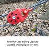 A red load-bearing snatch block is attached to a thick rope lying on a forest floor.