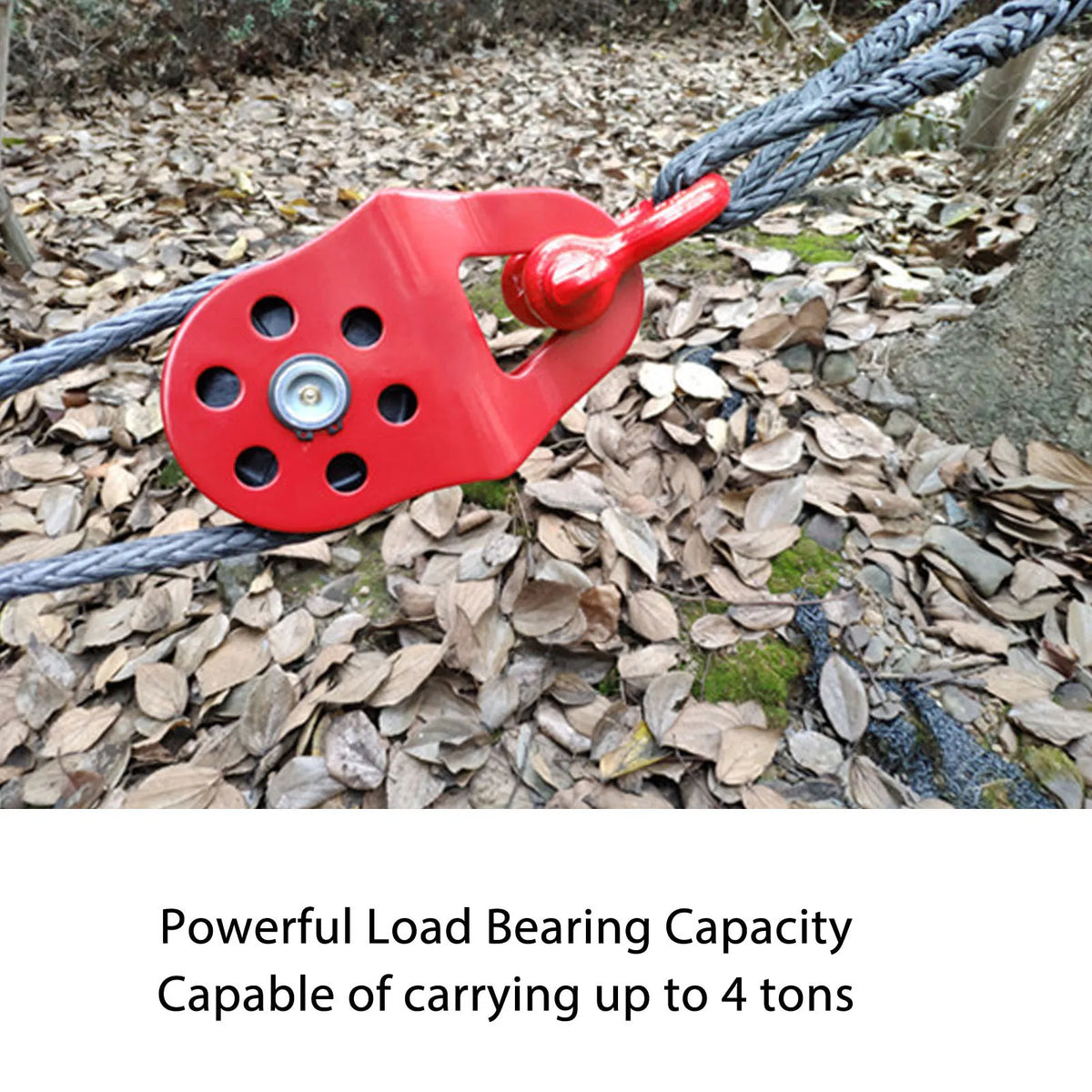 A red load-bearing snatch block is attached to a thick rope lying on a forest floor.