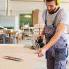 A man wearing safety glasses and an apron is using a precision contour gauge on a wooden surface in a workshop.