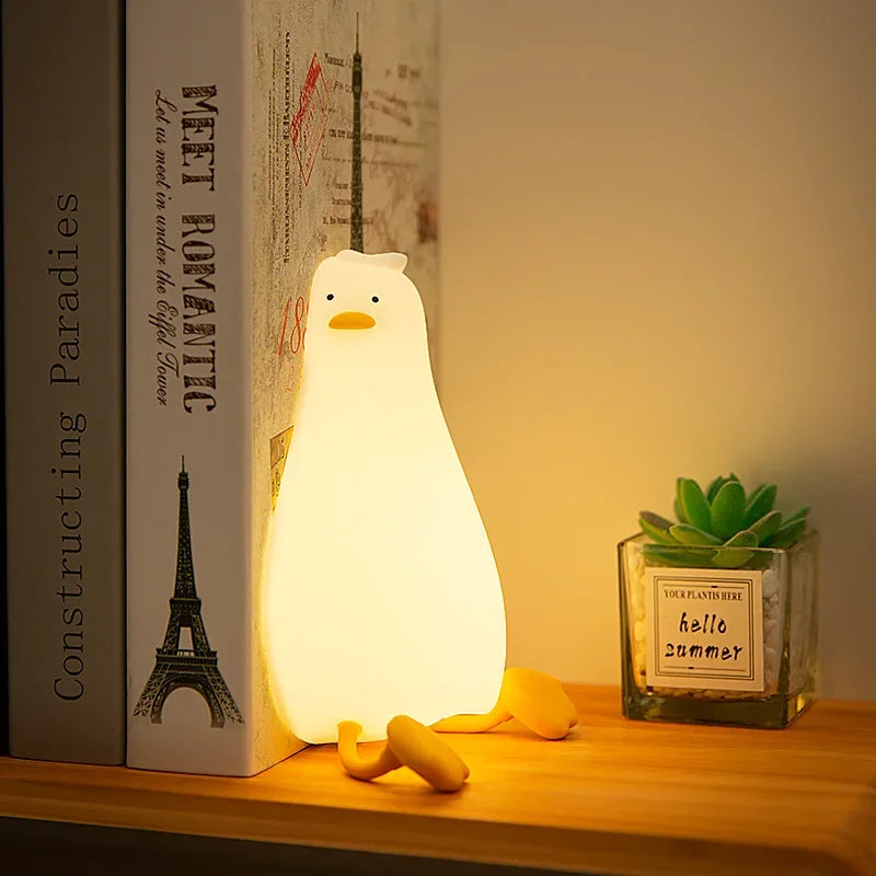 Weißer Silikon-Enten-Nachtlicht beleuchtet eine kleine Sukkulente auf einer Holzoberfläche neben einem Buch.