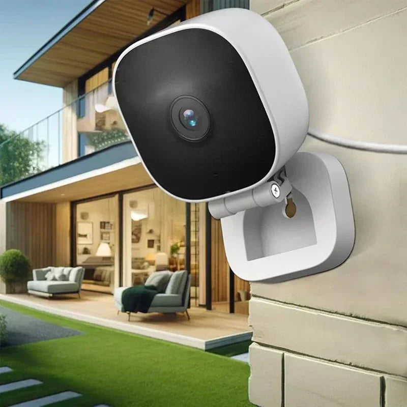 A white security camera is mounted on an exterior wall, overlooking a modern house and lawn.
