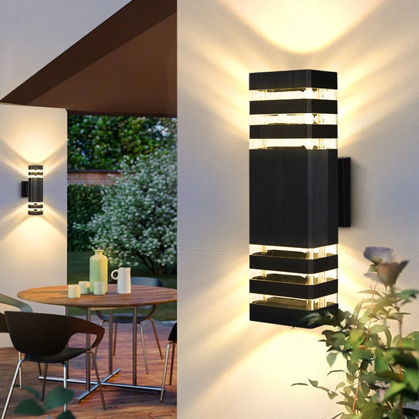 A modern black LED wall sconce with horizontal light strips illuminates a white wall outdoors.