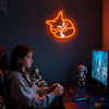 A young woman sits at a desk in a dimly lit room looking at an orange neon fox wall sign.