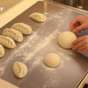 Hands are rolling dough balls on a stainless steel cutting board for baking and kneading.