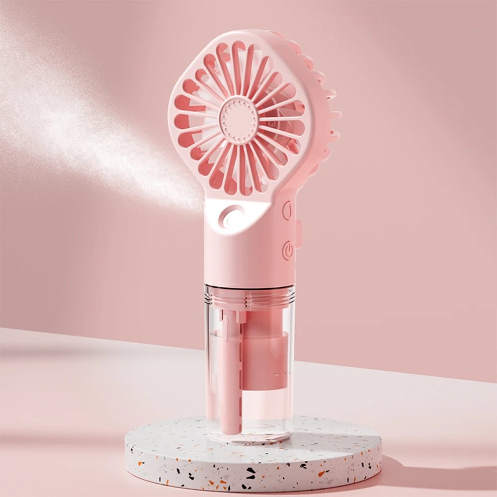 A pink portable USB rechargeable mist fan with a circular grille and a clear container is displayed on a white speckled surfa