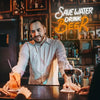 A man in a shirt stands in a dimly lit bar setting with an orange drink and a neon sign that reads Save Water Drink Beer