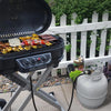A black grill is open, showing food, next to a propane tank pressure gauge and a white picket fence.
