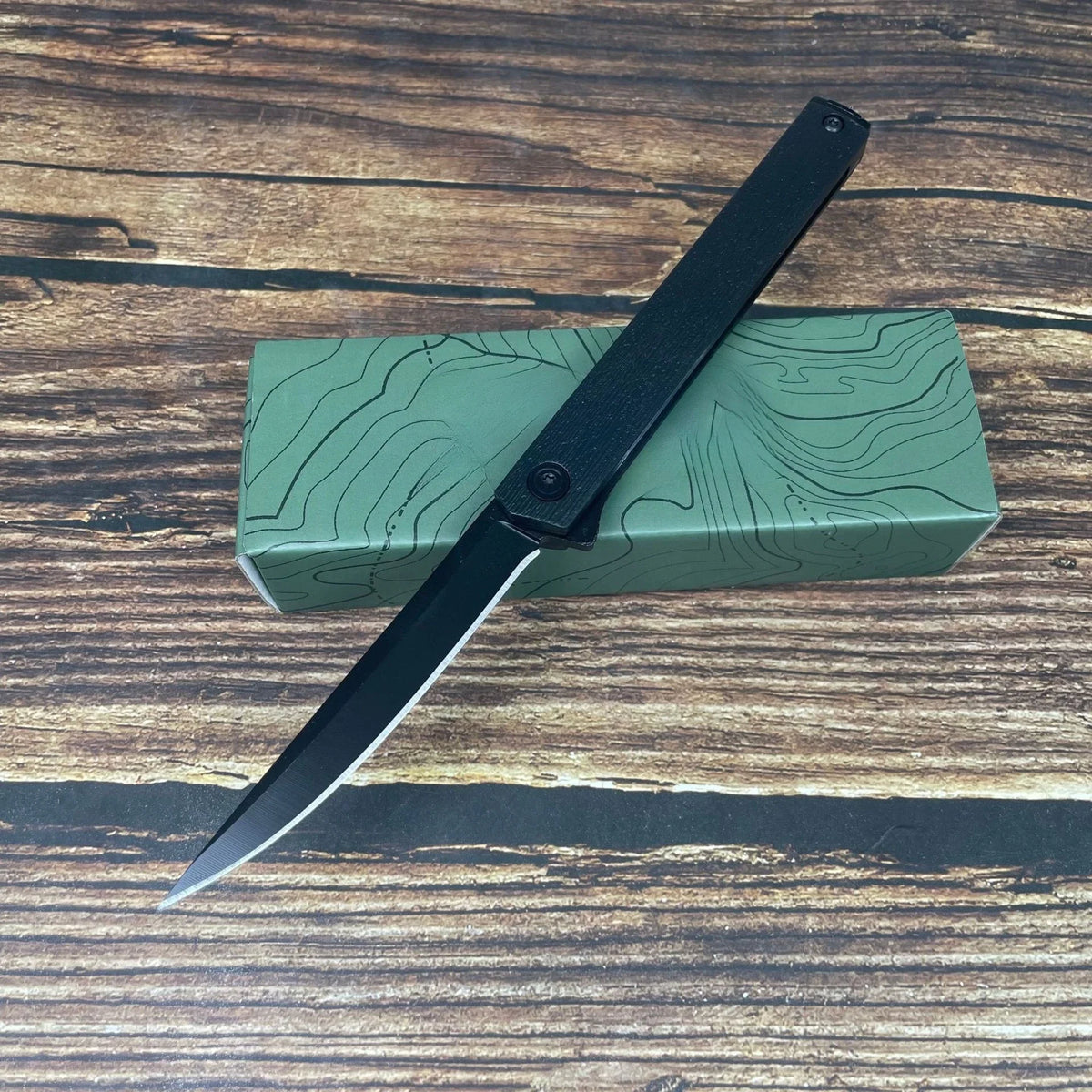 A black foldable utility knife with a sharp blade rests diagonally across a green wooden box.