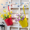 A realistic dragonfly clip in blue and silver is hanging on a white wooden panel next to potted yellow flowers.