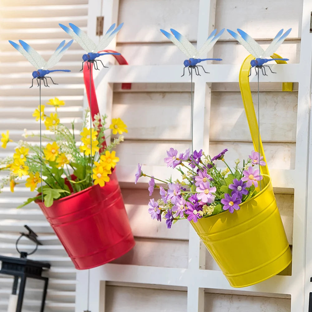 A realistic dragonfly clip in blue and silver is hanging on a white wooden panel next to potted yellow flowers.