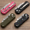 Three folding steel utility knives in pink, black, and olive green cases are displayed on a wooden surface.