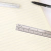 A silver aluminum triangular ruler is placed on a lined sheet of paper next to a black pen.