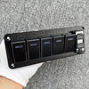 Panneau de commutateurs noir en aluminium avec cinq interrupteurs individuels et un port de charge, posé sur un tissu blanc.