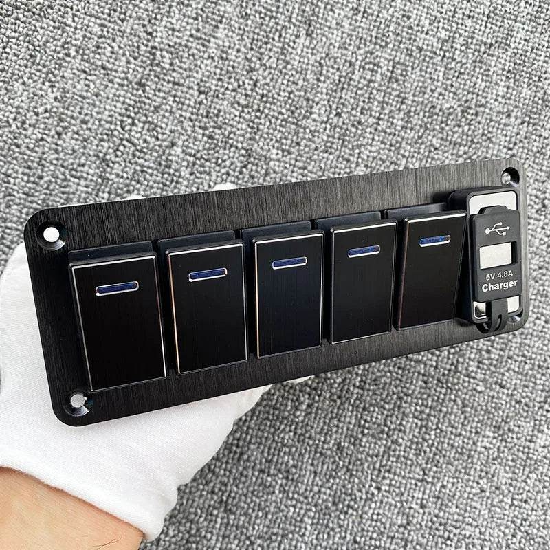 Panneau de commutateurs noir en aluminium avec cinq interrupteurs individuels et un port de charge, posé sur un tissu blanc.