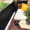 Una mesa de patio de madera al aire libre con cojines claros y almohadas amarillas junto a una pantalla negra de privacidad.