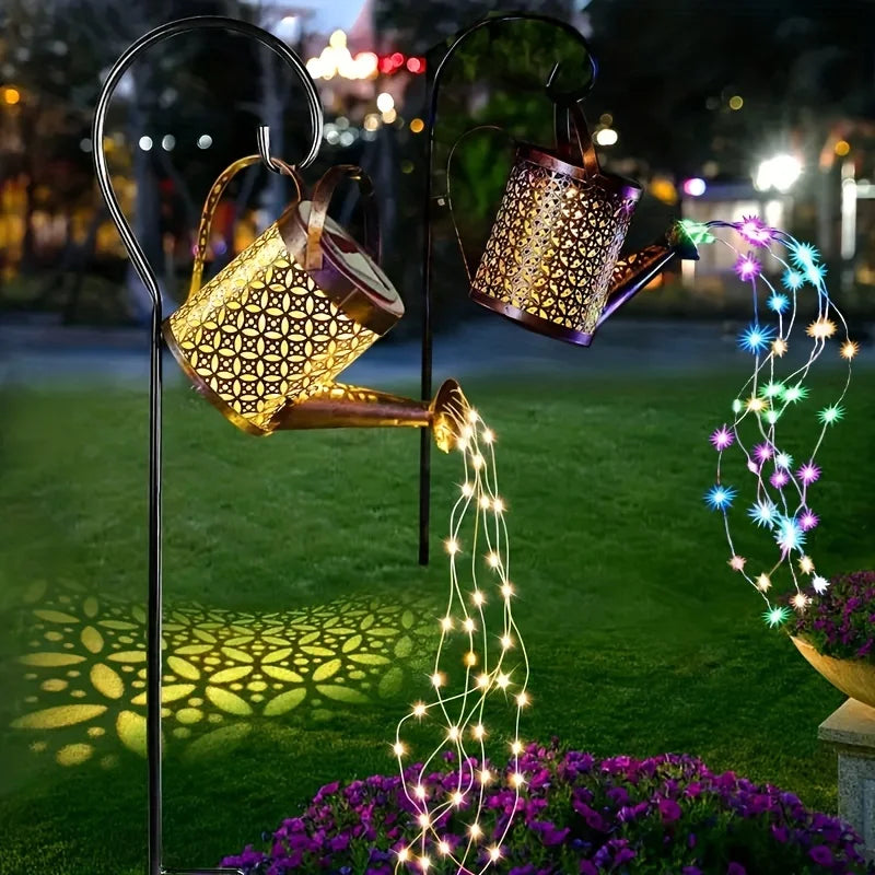 A solar fairy watering can light with glowing wires is displayed in a garden setting.