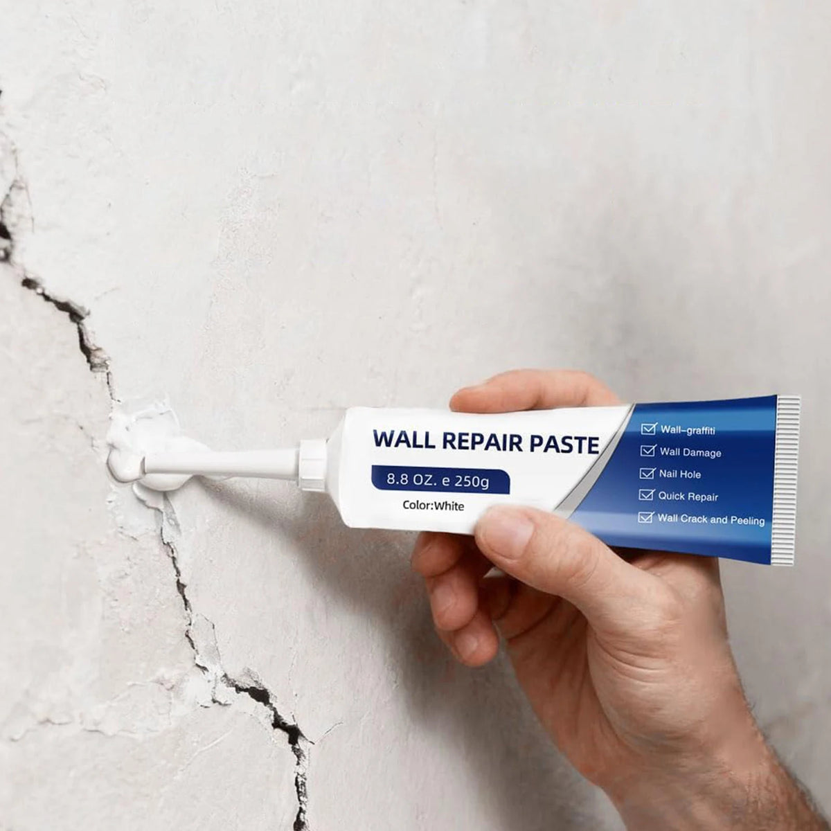 A hand holds a tube of Wall Repair Paste for drywall repair against a cracked white wall.