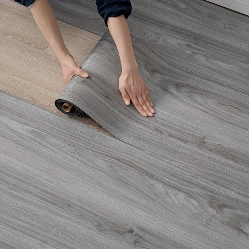 Hands are unrolling a roll of gray wood grain self-adhesive wall stickers onto a wooden floor.