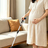 A woman holds a foldable magnetic reacher tool while standing next to a sofa and a small teddy bear on a light rug.