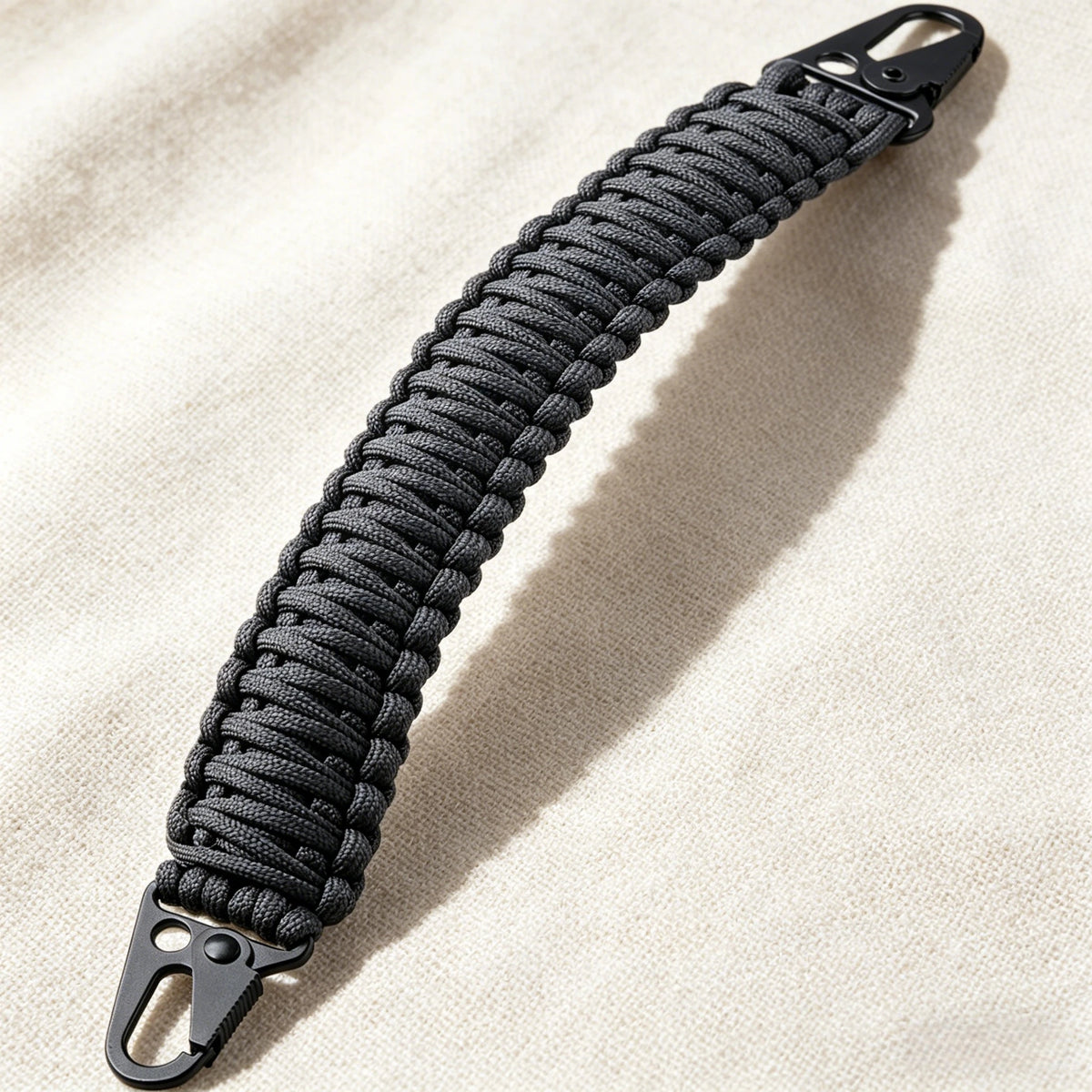 Schwarzer Paracord-Tragegurt mit Metallhaken vor hellbeigem Stoff.