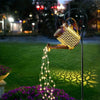 A solar fairy watering can light is shown illuminating a garden with glowing string lights.