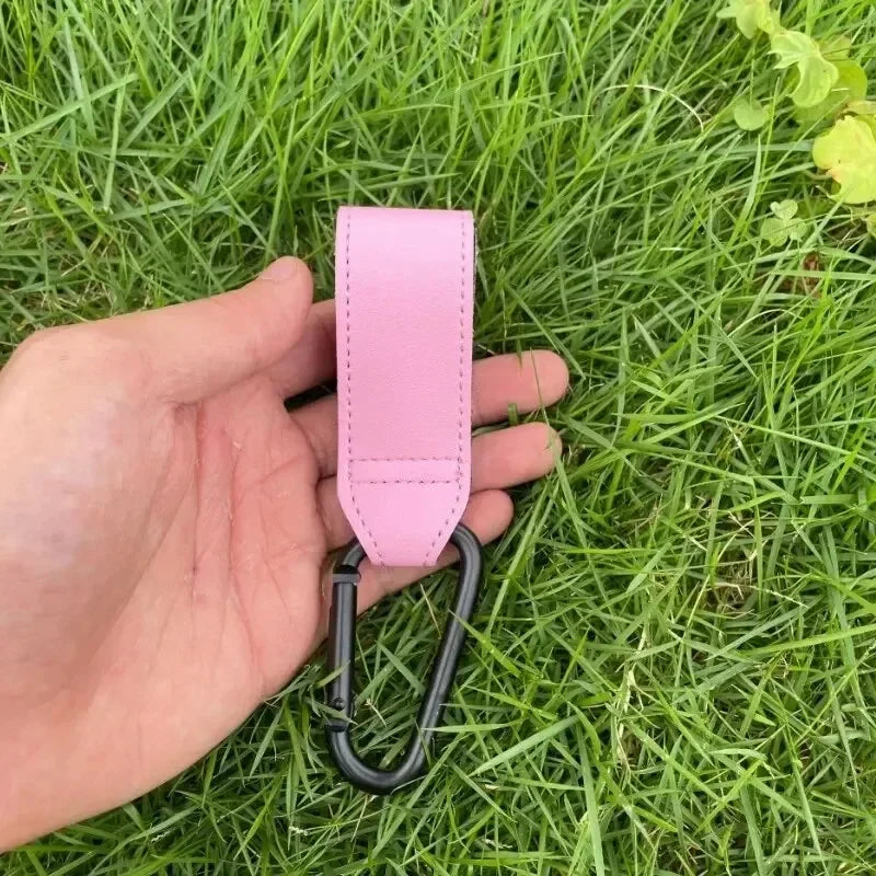 Une main tient une sangle en cuir rose avec un crochet, utilisée pour accessoires de poussette ou de sac à langer.