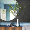 Jarrón decorativo con ramas verdes sobre una superficie oscura frente a una pared gris texturizada y zócal pintado de azul.