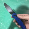 Une main tient un couteau EDC pliable bleu avec une lame noire sur fond vert.