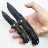 A hand holds a black folding survival knife with a textured handle and blade, displayed against a white background.