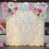 Weißer, drapierter Hintergrund mit rosa und cremefarbenen Blumenarrangements und beleuchtet von Lichterketten.