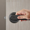 A hand holds a black smart fingerprint door lock device against a light wood grain door.