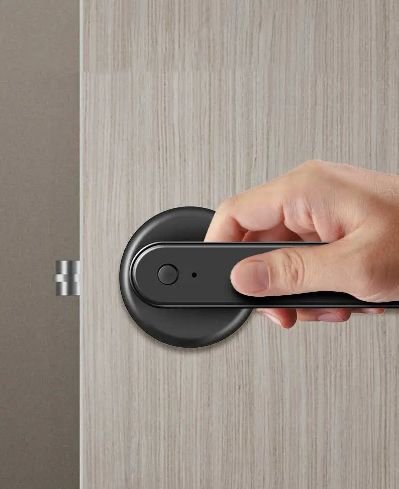 A hand holds a black smart fingerprint door lock device against a light wood grain door.