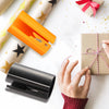 A hand is using a black sliding wrapping paper cutter next to a gold star-patterned paper and a wrapped gift.