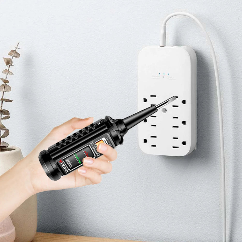 A hand holds a black pen with a tip, pointing toward a white AC voltage detection plug mounted on a wall.