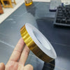 Eine Hand hält eine Rolle goldfarbigen selbstklebenden Fugenabdichtbandes auf einer grauen Oberfläche.