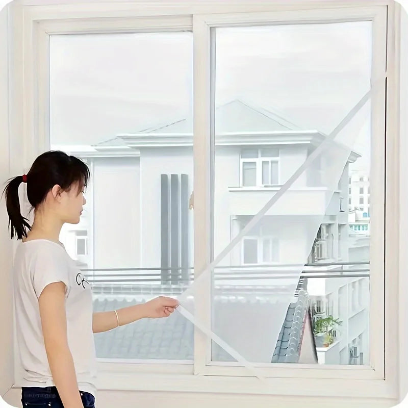 A woman is holding an invisible anti-mosquito window screen up to a large window overlooking a modern house.