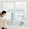 A woman is holding an invisible anti-mosquito window screen up to a large window overlooking a modern house.