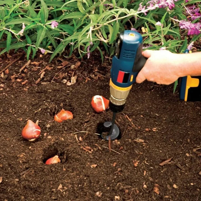 A hand is using a heavy-duty spiral garden auger drill bit to bore a hole in dark soil.