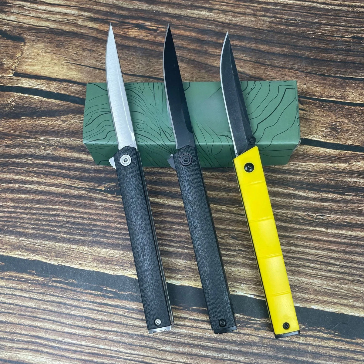 Three folding utility knives with black handles and silver blades resting on a green box on a wooden surface.