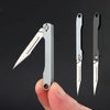 A hand holds a mini folding art knife, showing the blade and handle in profile against a dark background.