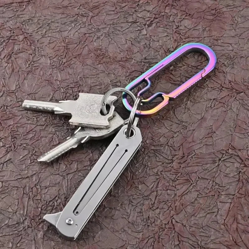 A stainless steel folding keychain knife with a silver blade and purple-colored keyring attached to a dark surface.